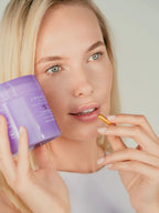 A close-up portrait of a blonde woman holding a purple Longevity Method supplement jar next to her face while holding a single gold capsule near her mouth